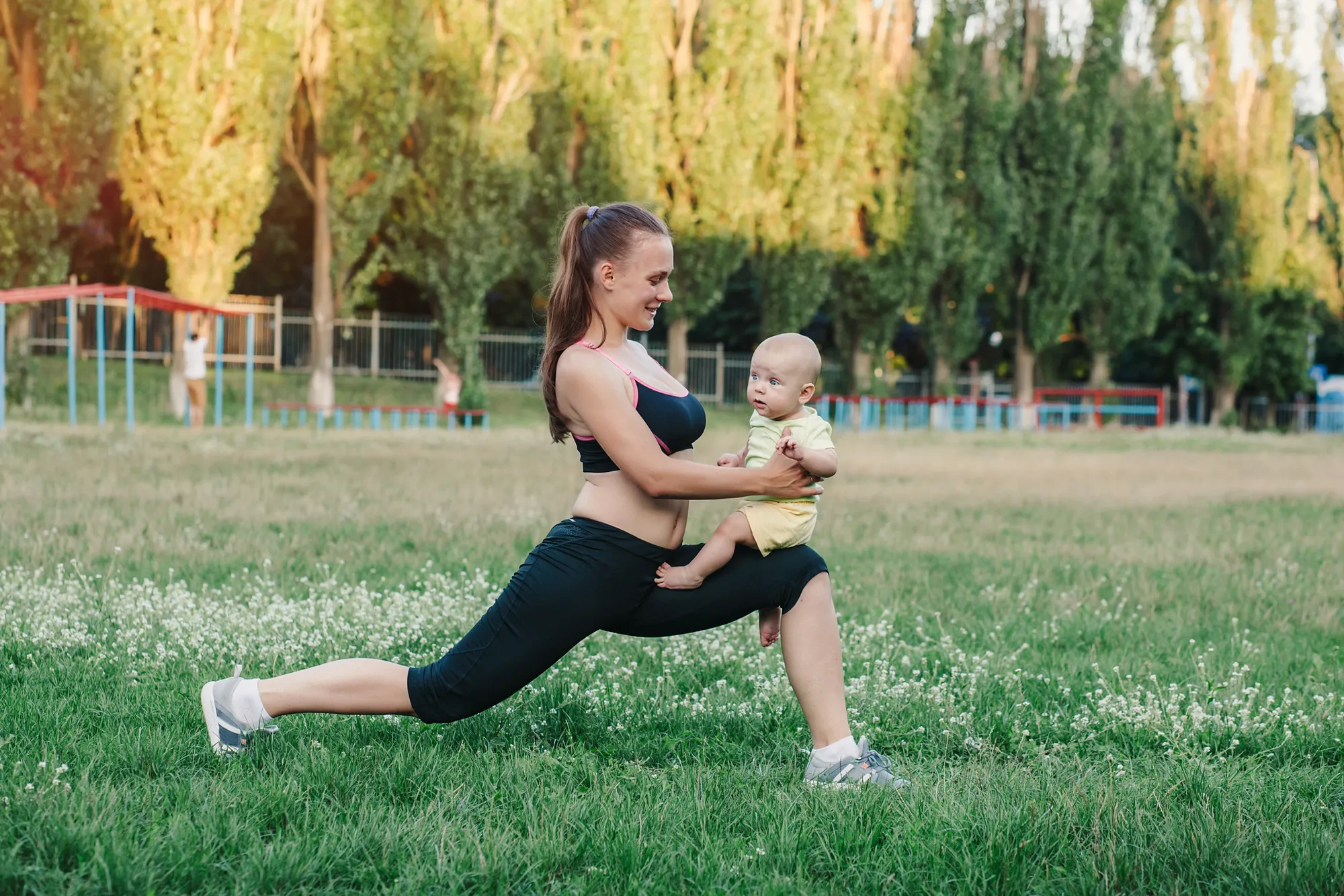 Frau in Sportkleidung macht im Park einen Ausfallschritt und hält dabei ihr Baby im Arm — sanfter, alltagsnaher Wiedereinstieg in den Sport nach der Geburt.