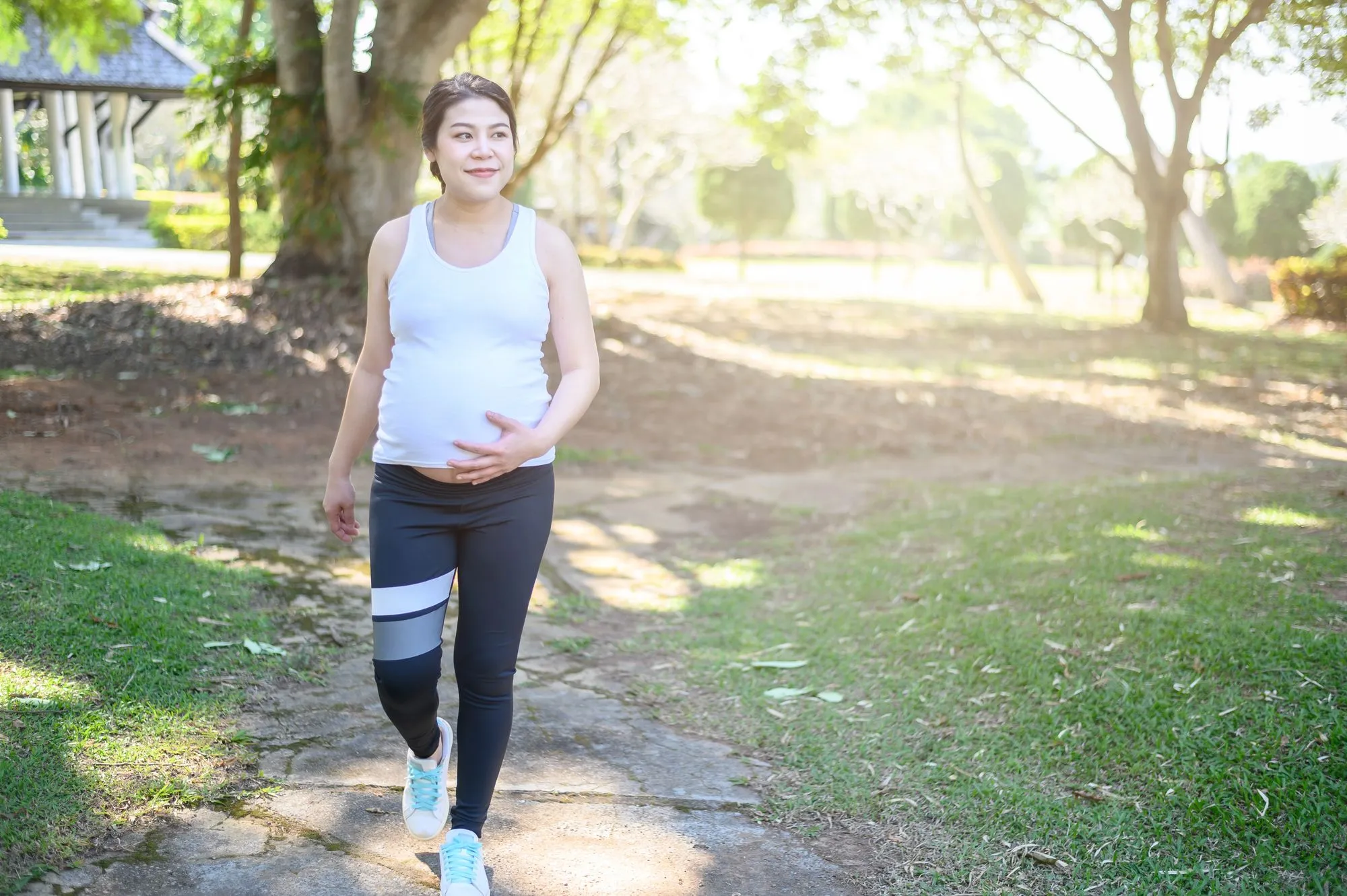 Schwangere Frau beim zügigen Spaziergang im Park, eine Hand am Babybauch