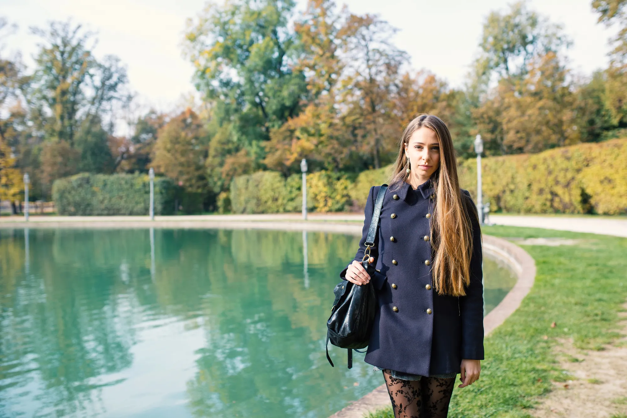 Frau im ersten Trimester geht an einem herbstlichen Parkteich spazieren, noch ohne sichtbaren Babybauch